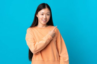 İzole edilmiş mavi arka planda genç bir Çinli kız bir ürünü sunmak için kenarı işaret ediyor.