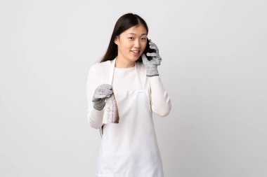 Önlük takan Çinli balıkçı, izole edilmiş beyaz arka planda çiğ balık tutuyor ve cep telefonuyla konuşan biriyle konuşuyor.