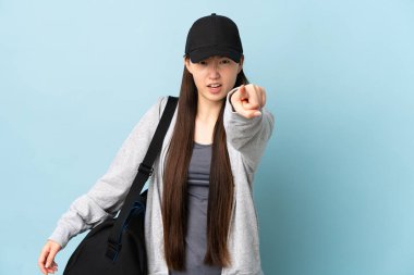 İzole edilmiş mavi arka planda spor çantalı genç Çinli kadın hayal kırıklığına uğramış ve ön tarafı işaret ediyor.