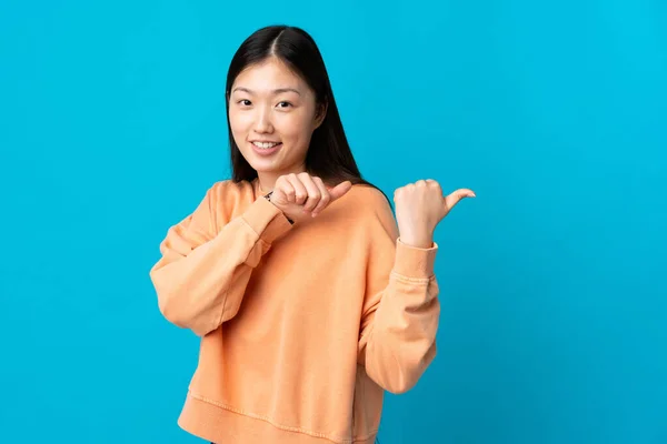 一位年轻的中国姑娘从孤立的蓝色背景中走出来 指着侧面展示自己的产品 — 图库照片