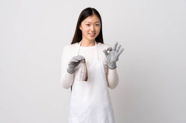 Çinli balıkçı önlük takıyor ve soyutlanmış beyaz arka planda çiğ balık tutuyor. Tamam işareti iki eliyle gösteriyor.