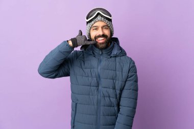 İzole edilmiş mor arka planda snowboard gözlüklü kayakçı adam telefon jesti yapıyor. Bana geri dönüş işareti ver.