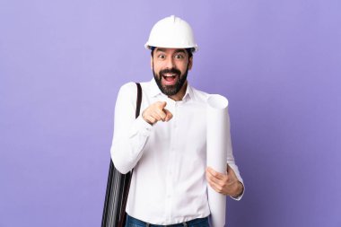 Miğferli genç bir mimar, izole edilmiş mor arka plan üzerinde planları var. Şaşırmış ve ön tarafı işaret ediyor.