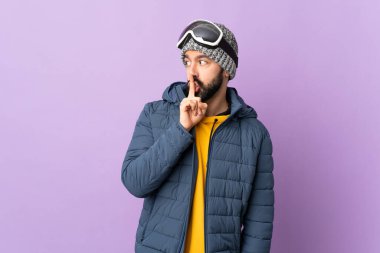 İzole edilmiş mor arka planda snowboard gözlüklü kayakçı parmağını ağzına koyan sessizlik işareti gösteriyor.