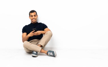 Yerde oturan Afrikalı Amerikalı adam cep telefonuyla mesaj yolluyor.