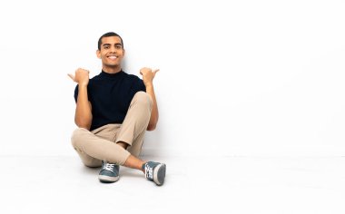 Afro-Amerikalı bir adam yerde oturuyor. Başparmakları havada ve gülümsüyor.