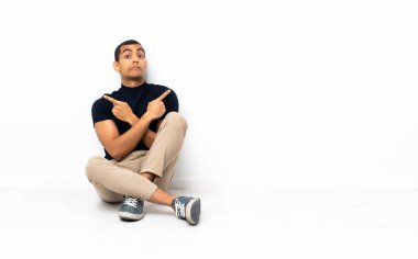 Afro-Amerikalı bir adam yerde oturuyor. Yan taraftakileri işaret ediyor. Şüpheleri var.