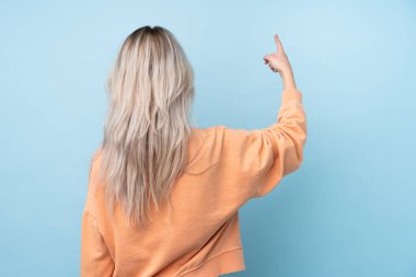 İzole edilmiş mavi arka planı olan genç kız işaret parmağıyla işaret ediyor.