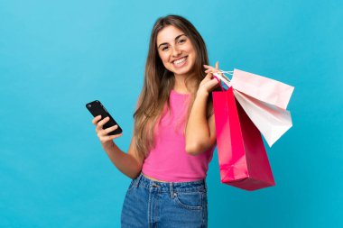Elinde alışveriş poşetleri olan ve cep telefonuyla bir arkadaşına mesaj yazan izole edilmiş mavi arka planda genç bir kadın.