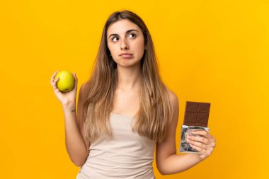 Bir elinde çikolata tableti, diğer elinde elma tutarken kuşkuları olan izole edilmiş sarı arka planda genç bir kadın.