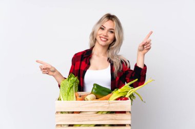 Bir sepet dolusu taze sebzeyi izole edilmiş beyaz arka planda tutan çiftçi kız parmak uçlarını işaret ediyor ve mutlu.