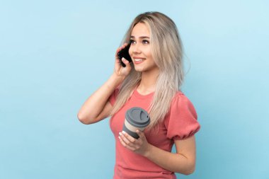 İzole edilmiş mavi arka planda, elinde kahve ve cep telefonu olan genç bir kız.