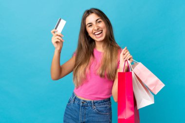 Soyutlanmış mavi arka planda genç bir kadın elinde alışveriş çantaları ve bir kredi kartı tutuyor.