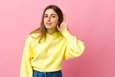 Soyutlanmış pembe arka planda genç bir kadın. Kulağına elini sürerek bir şeyler dinliyor.