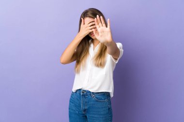 İzole edilmiş mor arka planda genç bir kadın dur hareketi yapıyor ve yüzünü kapatıyor.