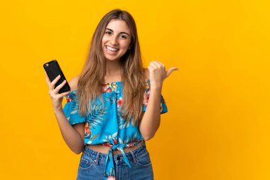 İzole edilmiş sarı arka planda cep telefonu kullanan genç bir kadın bir ürün sunmak için kenarı işaret ediyor.