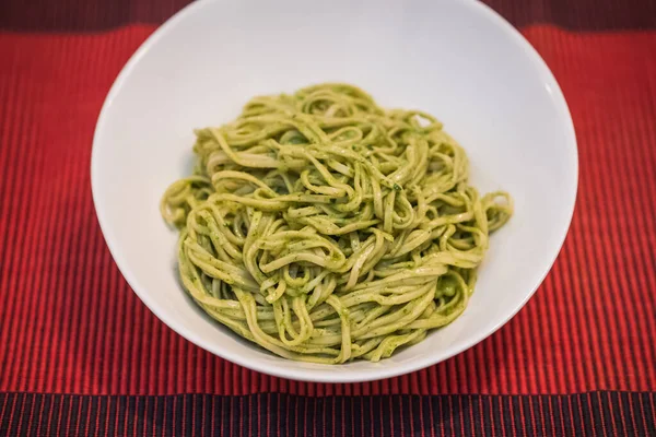 A white bowl of fresh egg pasta noodles with green pesto on a red background