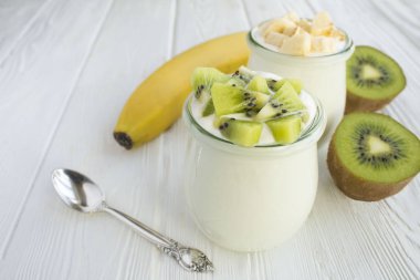 Beyaz ahşap arka planda cam kavanozlarda kivi ve muz bulunan doğal süt yoğurdu. Yakın çekim..