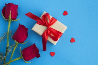 Kırmızı fiyonklu bir hediye, kırmızı güller ve mavi arka planda kırmızı kalpler. Üst Manzara. Boşluğu kopyala.