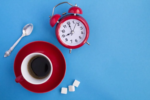 Coffee time. Composition with black coffee in the red cup and red alarm clock on the blue  background. Top view. Copy space.
