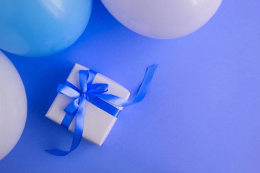 Mavi fiyonklu bir hediye, mavi arka planda mavi ve beyaz balonlar. Üst Manzara. Boşluğu kopyala.