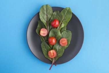 Mavi arkaplanın ortasındaki siyah tabakta marul ve vişneli domatesler. Üst Manzara. Boşluğu kopyala.