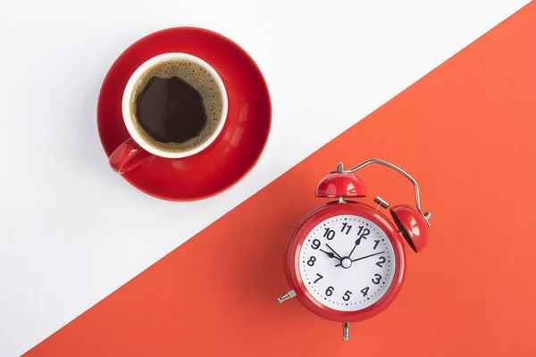 Coffee time. Composition with black coffee in the red cup and red alarm clock on the bicolor  background. Top view. Copy space.