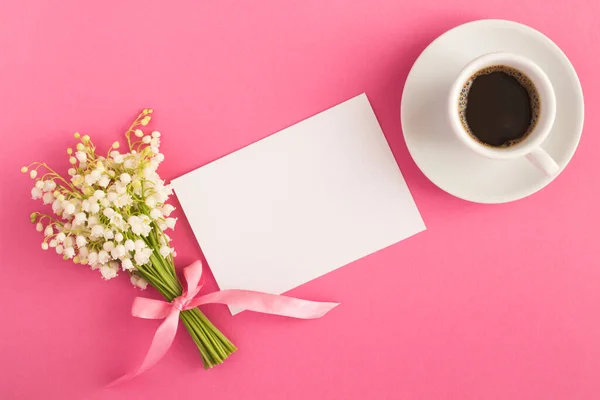 Blank white sheet of paper, black coffee in the  cup and bouquet of lilies of the valley on the pink  background. Top view. Copy space.