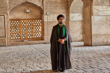 Isfahan, Iran - April 24, 2017: Iranian mullah in traditional clothes stands in the courtyard of the mosque. clipart