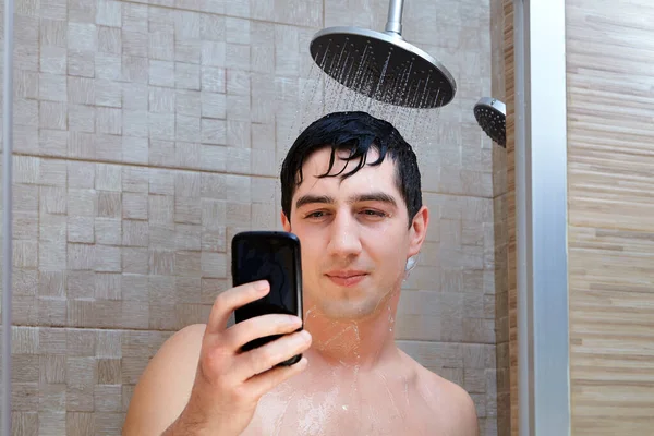 一位年轻貌美的白人年轻人站在从淋浴喷出的水柱下 一边在智能手机屏幕上读信息 湿淋淋的白人男子在浴室里用智能手机 — 图库照片