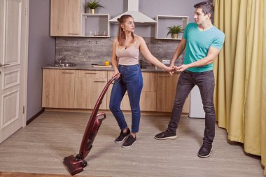 Çekici beyaz kadın mutfakta elektrikli süpürgeyle yerleri temizliyor ve erkek arkadaşı yakınlarda duruyor ve eline dokunuyor..
