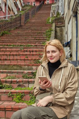 Genç beyaz bir kadın kışın ıssız İstanbul 'da tek başına yürüyor, sıcak kıyafetler giyiyor ve dinleniyor, akıllı telefonuyla büyük kırmızı bir merdivenin arka planında haritayı inceliyor..