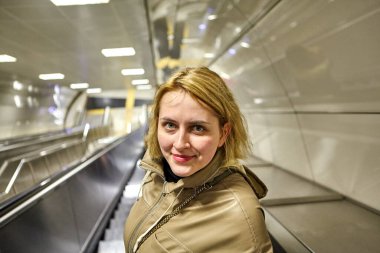 İstanbul metrosundaki yürüyen merdivende, günlük yağmurluk giymiş, oldukça neşeli, beyaz bir kadın duruyor..