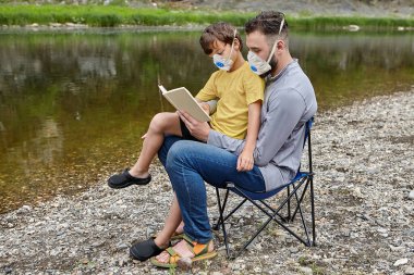 Fathers Day during quarantine in connection with the covid-19 pandemic, father and son use medical facemasks to protect against infection. Young man and child in N95 respirator masks near river. clipart