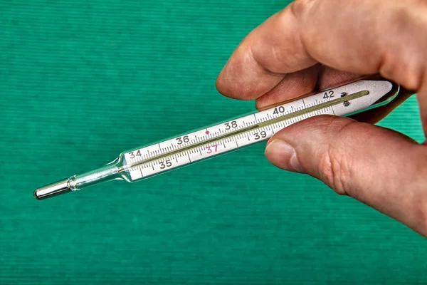 A glass mercury thermometer shows the high temperature of the human body on its scale. Man\'s hand holds a diagnostic medical tool or measuring device, on a green background.