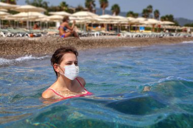 Genç Kafkasyalı kadın güneşli yaz günlerinde plaj şemsiyeleriyle kıyı şeridinin arka planında koruyucu tıbbi maskeyle denizde yüzüyor. Coronavirüs enfeksiyonu salgınında tatiller Covid-19.