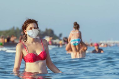 Plaj sezonu salgın döneminde, Kovid-19. Tatil sırasında genç bir kadın denizde banyo yapar. Beyaz kadın koruyucu bir yüz maskesi takıyor..