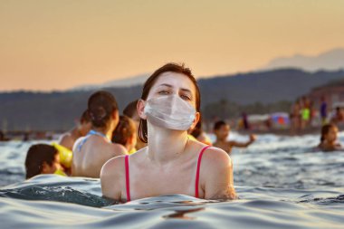 Deniz kıyısındaki bir tatil yerinde Coronavirus enfeksiyonuna karşı korunma. Kovid-19 salgını sırasındaki sahil sezonunda genç bir Kafkas kadın koruyucu bir maskeyle denizde yıkanır..