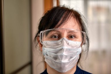 Orta yaşlı bir kadın içeride maske ve güvenlik gözlüğü takıyor. Beyaz dişi, karantina sırasında koronavirüs enfeksiyonu Covid-19 salgınıyla ilgili olarak PPE kullanıyor..