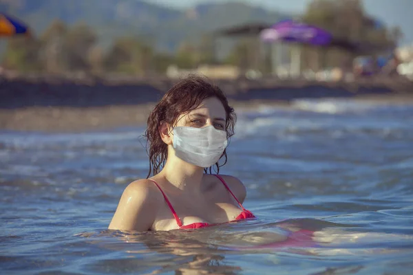 Young woman is swimming in the water of the sea on resort with medical facial disposable mask for respiratory protection on her face during coronavirus infection covid-19 pandemic and home isolation.