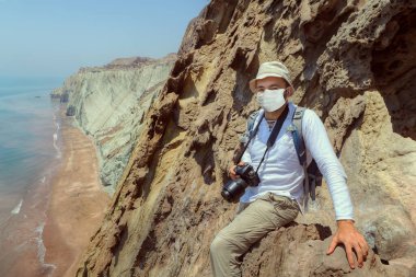 Turizm sezonu 2020 Hormozgan Eyaleti Hormuz Adası, İran 'ın güneyinde. Koruyucu sağlık maskeli beyaz gezgin elinde kamerayla bir kayanın üzerinde oturuyor..