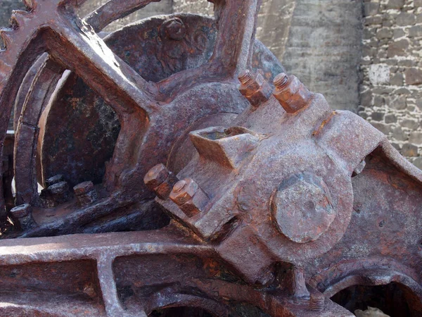 Close up of an axle and broken spoked wheel on old rusted abandoned industrial machinery against a stone wall — Stock Photo, Image