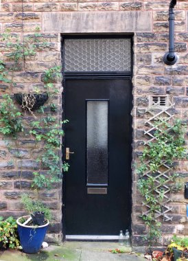 a black door in a typical traditional yorkshire house with stone walls surrounded by plants in pots and milk bottles