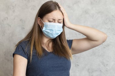 Tıbbi maske takan mutsuz genç bir kadının başı ağrıyor. Kız acı çekiyor. Soğuk algınlığının belirtileri. Grip veya virüs. Evde migreni olan yorgun bir kız..