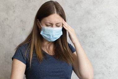 Tıbbi maske takan mutsuz genç bir kadının başı ağrıyor. Kız acı çekiyor. Soğuk algınlığının belirtileri. Grip veya virüs. Evde migreni olan yorgun bir kız..