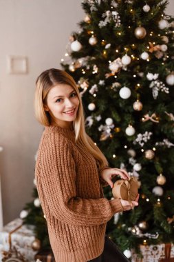 Kahverengi örgü süveterli sarışın elinde hediyesini elinde tutan süslü bir Noel ağacının yanında. Noel fotoğrafı