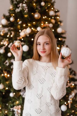 Mavi gözlü güzel sarışın kız elinde beyaz süveter giymiş bir Noel topunu Yeni Yıl ağacının arka planında tutuyor. Yılbaşı fotoğrafları. Noel fotoğrafı