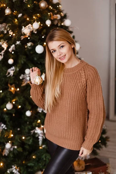 Rubia en un suéter de punto marrón sostiene un regalo en sus manos junto a un árbol de Navidad decorado, sosteniendo presente. Foto de Navidad —  Fotos de Stock