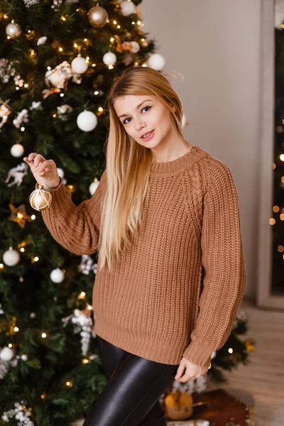Bionda in una maglia marrone maglione tiene un regalo tra le mani accanto a un albero di Natale decorato, tenendo presente. Foto di Natale — Foto Stock