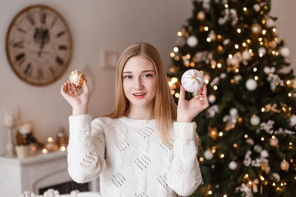 Piękna blondynka z niebieskimi oczami trzyma w dłoniach świąteczną kulę w białym swetrze na tle noworocznej choinki. Zdjęcia noworoczne. Zdjęcie świąteczne — Zdjęcie stockowe